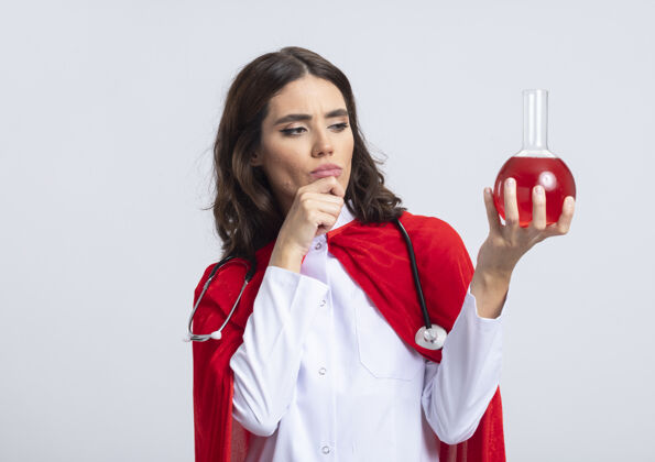 液体自信的女超人穿着医生制服 戴着红色斗篷和听诊器 把手放在下巴上 看着隔离在白墙上的玻璃瓶中的红色化学液体听诊器超级英雄瓶子