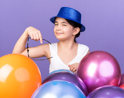 抱着恼怒的年轻白种女孩戴着蓝色的聚会帽 手持光学眼镜 站在紫色的墙上 氦气球与复制空间隔离恼火气球光学