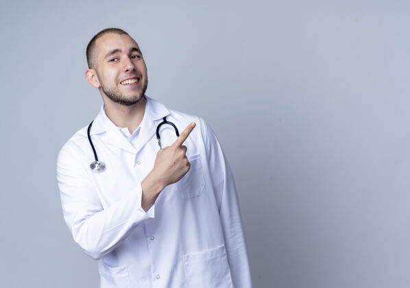 医生面带微笑的年轻男医生 身穿医用长袍 脖子上戴着听诊器 指着隔离在白墙上的一侧表情男性姿势