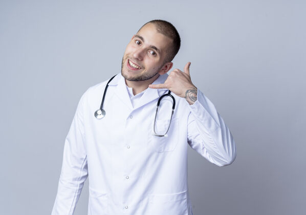 人面带微笑的年轻男医生穿着医用长袍 脖子上戴着听诊器 在白墙上做着孤立的呼叫手势听诊器衣服表情
