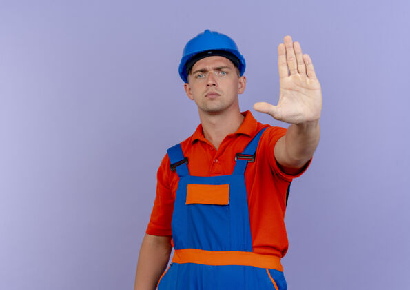 显示严格的年轻男性建筑工人穿着制服和安全帽显示停止手势男性头盔安全