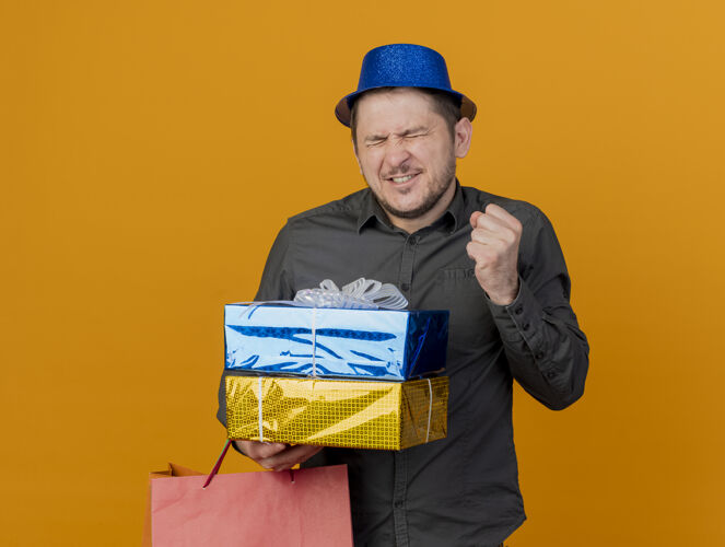帽子快乐的年轻人闭着眼睛戴着蓝色的帽子 拿着礼品盒和袋子 在橙色上显示“是”的手势包橙色显示