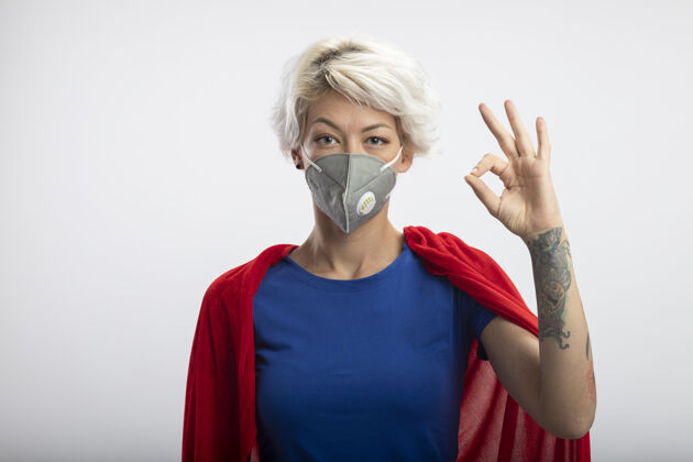 医学自信的女超人穿着红色斗篷戴着医用面罩手势ok手势隔离在白墙上公民面具女人