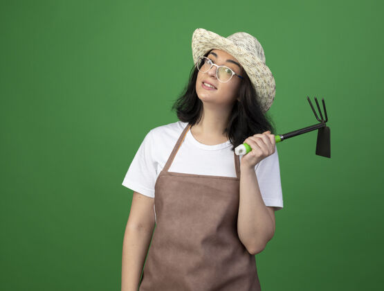 人令人印象深刻的年轻黑发女园丁在光学眼镜和制服戴园艺帽子举行锄头耙隔离在绿色的墙壁上锄头姿势脸