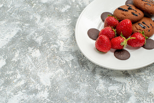 草莓上半部分查看巧克力饼干草莓和圆形巧克力在白色椭圆形盘子在顶部的灰白色地面右上角磨碎的可食用水果水果