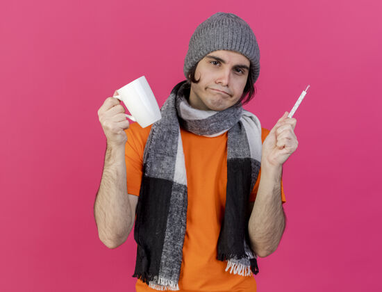 围巾未出狱的年轻病夫戴着冬天的帽子 戴着围巾 手里拿着一杯茶 温度计被隔离在粉色上茶体温计粉色
