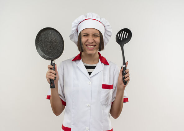 举行面带微笑的年轻女厨师 身着厨师制服 拿着抹刀和煎锅 闭着眼睛站在白色的墙上白微笑表情