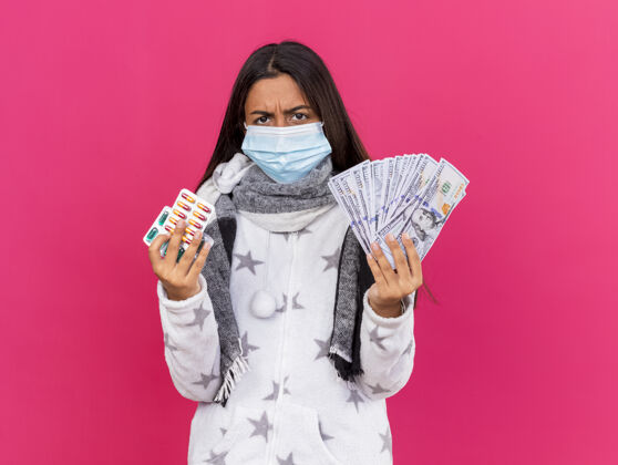 现金愤怒的年轻生病的女孩戴着医用面罩 戴着围巾 手里拿着药片 身上隔离着粉红色的现金年轻药片粉色
