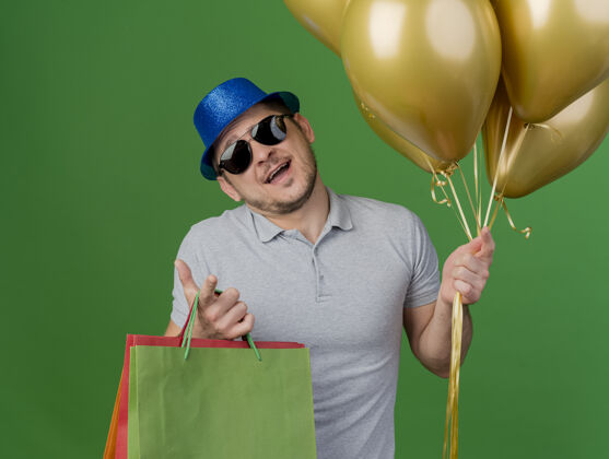 礼物一个戴着派对帽和眼镜 拿着气球和礼品袋的年轻人被隔离在绿色地带小伙子穿聚会