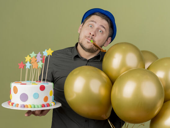 年轻困惑的年轻人穿着黑色衬衫 戴着蓝色帽子 拿着蛋糕和气球 吹着橄榄绿的派对吹风机穿蓝色气球