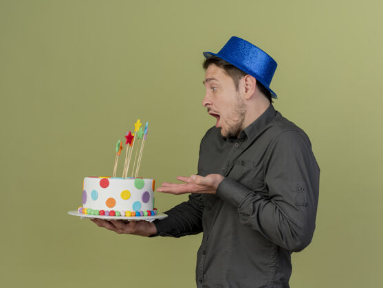 黑色站在侧视图惊讶的年轻人穿着黑色衬衫和蓝色的帽子党举行的手指着蛋糕孤立在橄榄绿穿年轻绿色