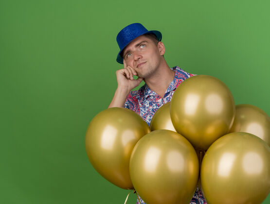 年轻体贴的年轻人戴着蓝色帽子站在气球后面 把手放在绿色的脸颊上思考站气球