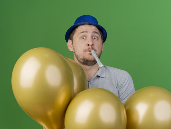 帽子惊讶的年轻人戴着派对帽站在气球后面吹派对风机在绿色年轻小伙子站