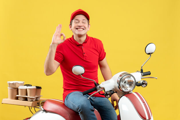成人前视图自信的年轻人穿着红色上衣 戴着帽子 在黄色背景下传递订单 做眼镜手势轻便摩托车车辆帽子