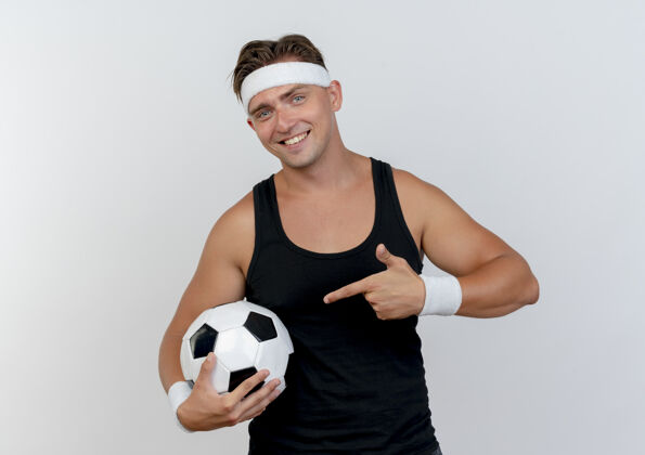 男人面带微笑的年轻英俊的运动型男子戴着头带和腕带拿着并指着隔离在白墙上的足球市民脸人