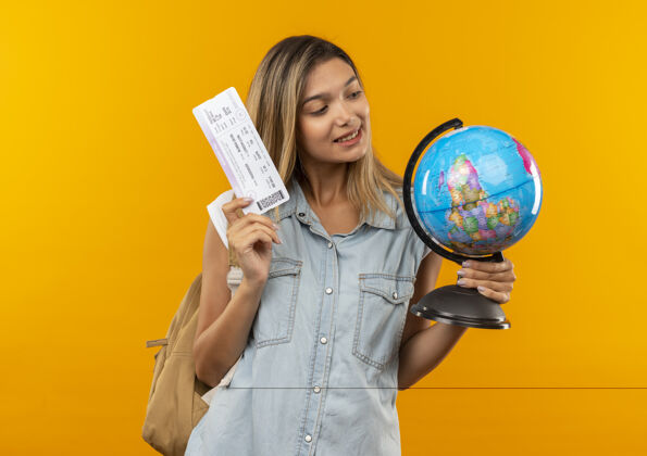 表情高兴的年轻漂亮的学生女孩戴着背包拿着地球仪和机票看着地球仪孤立在橙色的墙上公民脸包