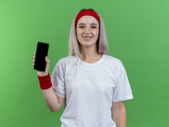 背带微笑的年轻运动女性戴着背带 戴着头带和腕带 把手机孤立在绿色的墙上运动女孩人