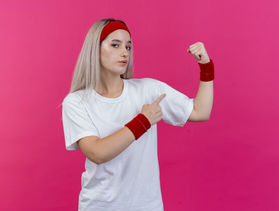 女人自信的年轻运动女性 戴着背带 戴着头带和腕带 紧绷着 指着隔离在粉色墙上的二头肌二头肌点紧张