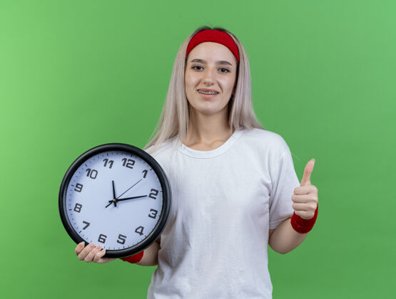 拇指微笑的年轻运动女性戴着背带 戴着头带和腕带 把时钟和大拇指孤立地竖立在绿色的墙上年轻时钟女人