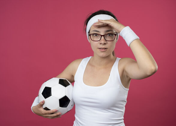 腕带戴着眼镜 戴着头带和腕带的快乐的年轻运动女性 手掌放在前额 把球孤立地举在粉红色的墙上前额手掌年轻