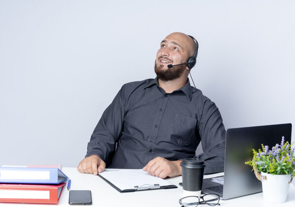 坐着微笑着的年轻秃头呼叫中心男子戴着耳机坐在办公桌旁 带着工作工具孤立地仰望着白色的墙壁工具表情人