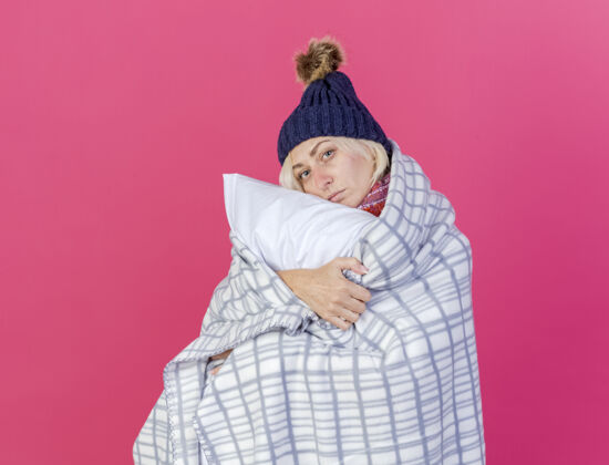 斯拉夫自信的金发生病的年轻女人戴着冬天的帽子 围巾裹着格子呢 抱着一个孤立在粉红色墙上的枕头女人疾病年轻人