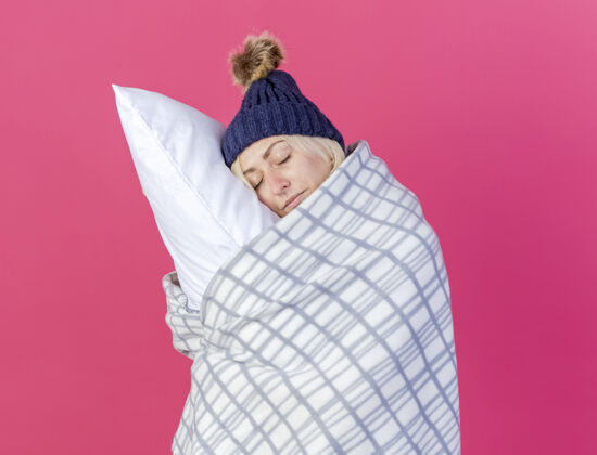 人昏昏欲睡的年轻金发病女人戴着冬天的帽子和围巾 裹着格子呢的拥抱 把头放在隔离在粉红色墙上的枕头上拥抱头围巾