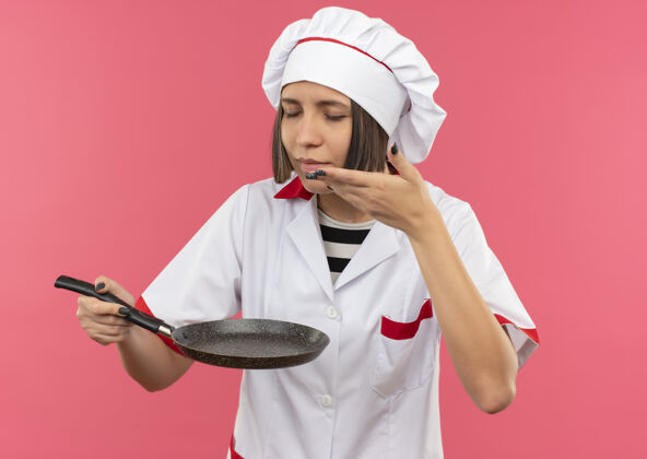 公民身着厨师制服的年轻女厨师手拿煎锅 闭着眼睛闻着 手放在粉色墙壁上的空气中人空气衣服
