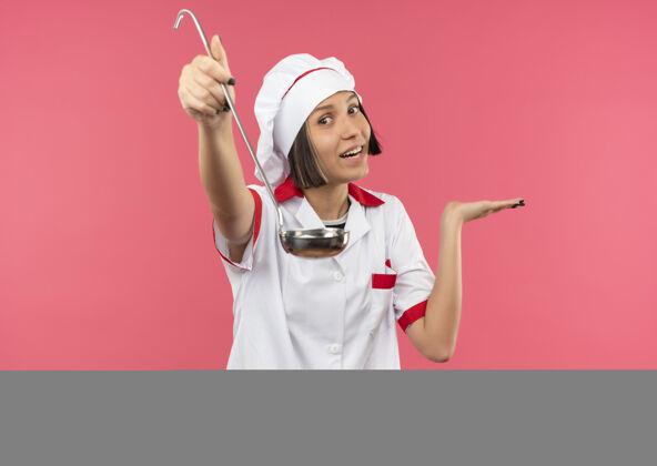 女性身着厨师制服的年轻女厨师微笑着伸出勺子 在粉红色的墙上孤立着一只空手表演衣服烹饪