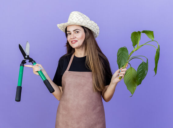 花园微笑美丽的园丁女孩穿着制服 戴着园艺帽 拿着剪子的植物 摊开双手孤立在蓝色上穿举行蓝色