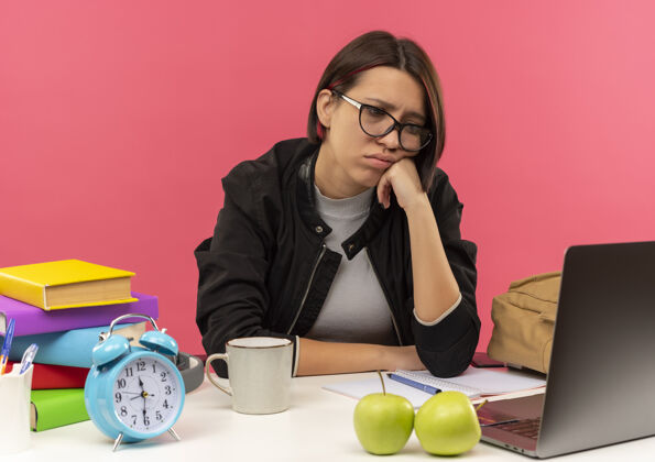 粉色戴着眼镜坐在书桌旁做作业的悲伤的年轻学生女孩把手放在脸上看着隔离在粉红色墙上的笔记本电脑人人表情