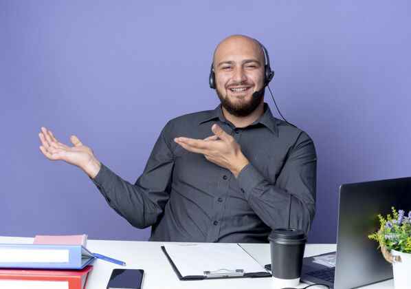 指向微笑着的年轻秃头呼叫中心男子戴着耳机坐在办公桌旁 手里拿着工作工具 伸出空手 指着紫色墙上孤立的那只手人感觉展示
