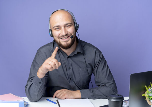 人微笑着的年轻秃头呼叫中心男子戴着耳机坐在办公桌旁与工作工具举起手指 把手放在桌子上孤立在紫色的墙上手指人手