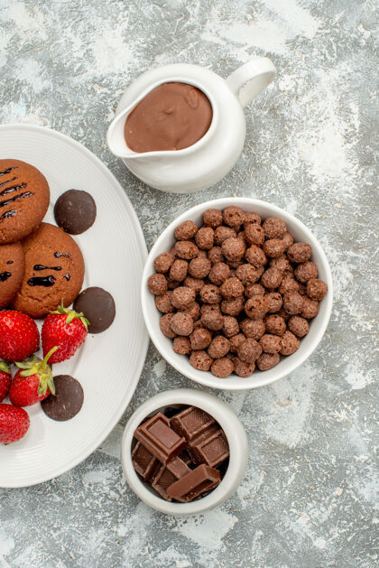 小吃白色椭圆形盘子上的巧克力饼干草莓和圆形巧克力 灰白色地面上的巧克力谷类和可可碗草莓碗谷类食品