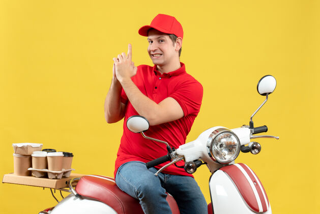 微笑顶视图自信的年轻人穿着红色上衣 戴着帽子 在黄色背景上做着枪的手势 传达着命令摩托车年轻人黄色