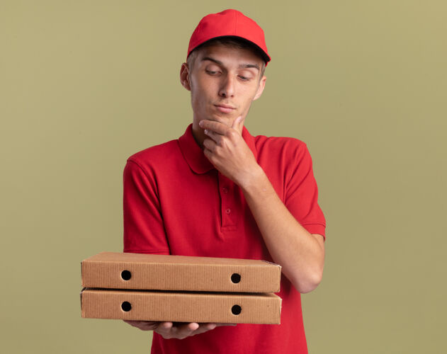 金发年轻的金发送货员高兴地把手放在下巴上拿着 看着橄榄绿的比萨饼盒下巴送货男孩
