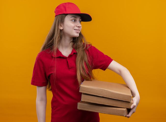 微笑在孤立的橙色空间里 身着红色制服 手持包裹 面带微笑的年轻送货员看着右边女孩制服右边
