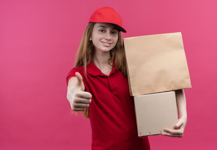 展示带着微笑的年轻送货女孩 穿着红色制服 拿着盒子 在有复印空间的粉色空间里竖起大拇指递送向上粉色