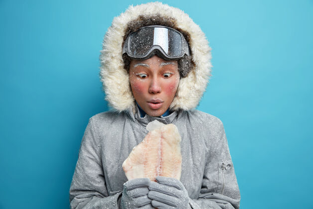 年轻震惊尴尬的美国黑人妇女盯着冻鱼穿着暖和的衣服感觉舒服戴着滑雪镜享受寒假在好时光感觉寒冷娱乐钓鱼奇迹