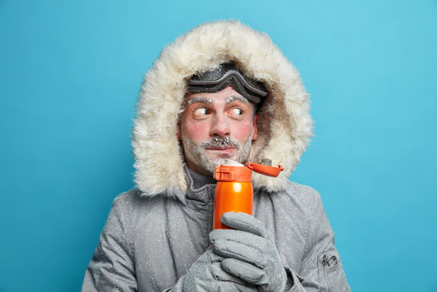 蓝色惊讶的留着胡须的欧洲男人满身冰饮料热饮料拿着保温瓶戴着滑雪护目镜和保温夹克人抱着夹克