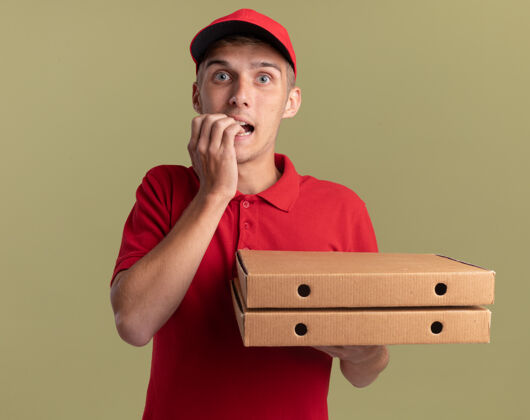 男孩焦急的年轻金发送货员咬指甲 拿着橄榄绿的比萨盒焦虑咬金发