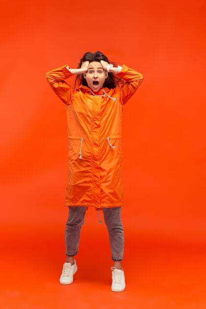 聪明这个快乐微笑的年轻女孩穿着秋天的橘色夹克在摄影棚里摆姿势红色隔离人类积极的情感寒冷天气的概念女性时尚概念下雨情绪冻结