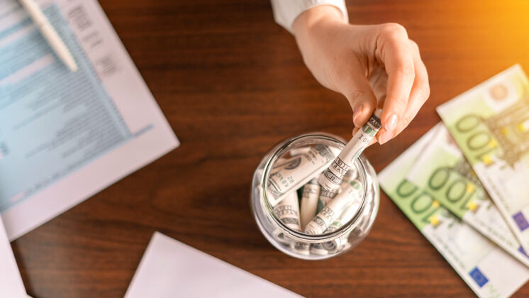 投资一个女人把钞票扔进一个罐子里 桌子上放着卷好的钞票纸 钱在桌子上资本粉色盒子