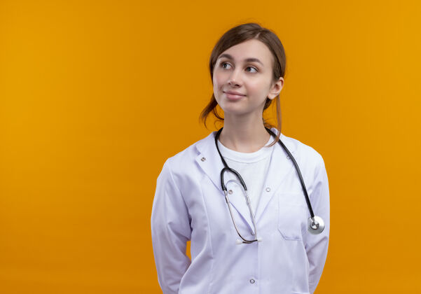 复制高兴的年轻女医生穿着医用长袍和听诊器 在孤立的橙色空间与复印空间左侧看医疗太空请穿