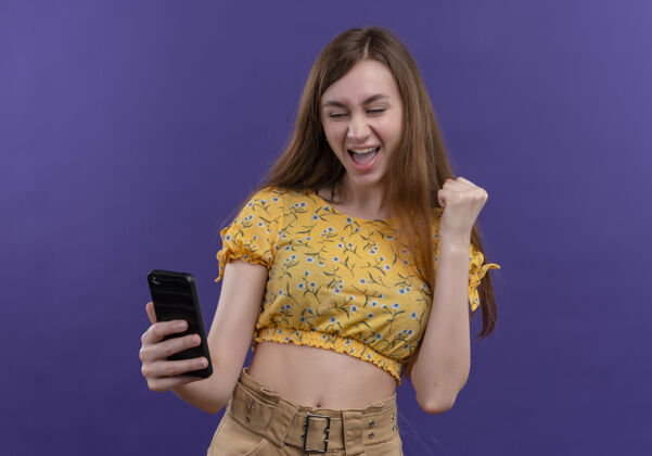 年轻快乐的年轻女孩拿着手机 在孤立的紫色空间举起拳头女孩手机快乐
