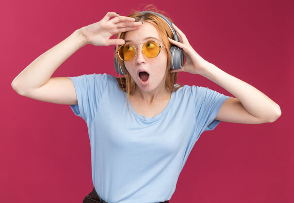 印象令人印象深刻的年轻红发姜女雀斑在太阳眼镜和耳机保持手掌在额头看着粉红色的一面保持太阳耳机
