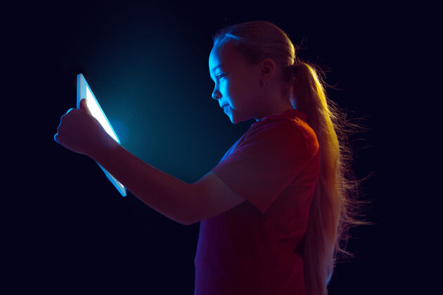 情绪在霓虹灯下 白种人女孩的肖像被隔离在黑暗的工作室背景上漂亮的女模特使用平板电脑人类情感的概念 面部表情 销售 广告 现代科技 小玩意使用虚拟日志衬衫