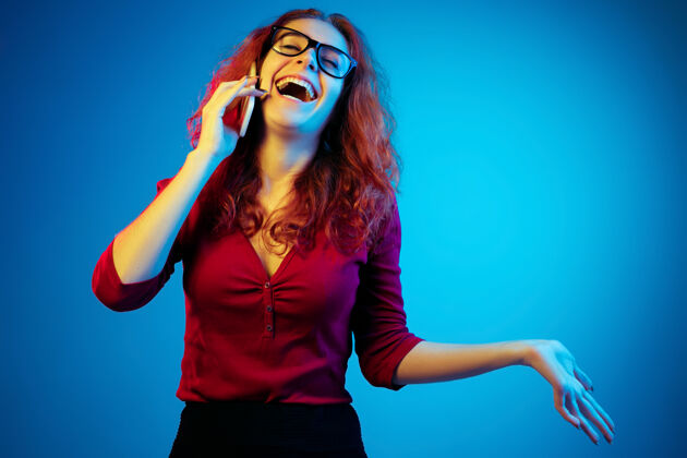 工作在霓虹灯下的蓝色工作室背景上孤立的高加索女性肖像随意的红色头发的美丽女性模特人类情感的概念 面部表情 销售 广告电话交谈衬衫执行成功