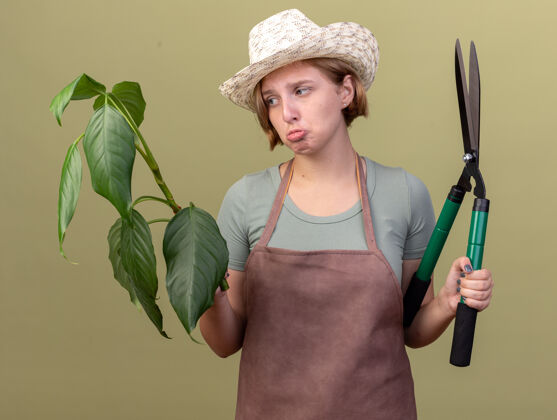 帽子悲伤的年轻斯拉夫女园丁戴着园艺帽手持园艺剪刀 在橄榄绿上种植斯拉夫剪刀橄榄