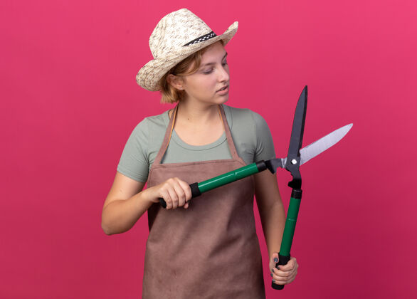 年轻的自信的年轻斯拉夫女园丁戴着园艺帽拿着园艺剪刀看着粉色女的园艺花园